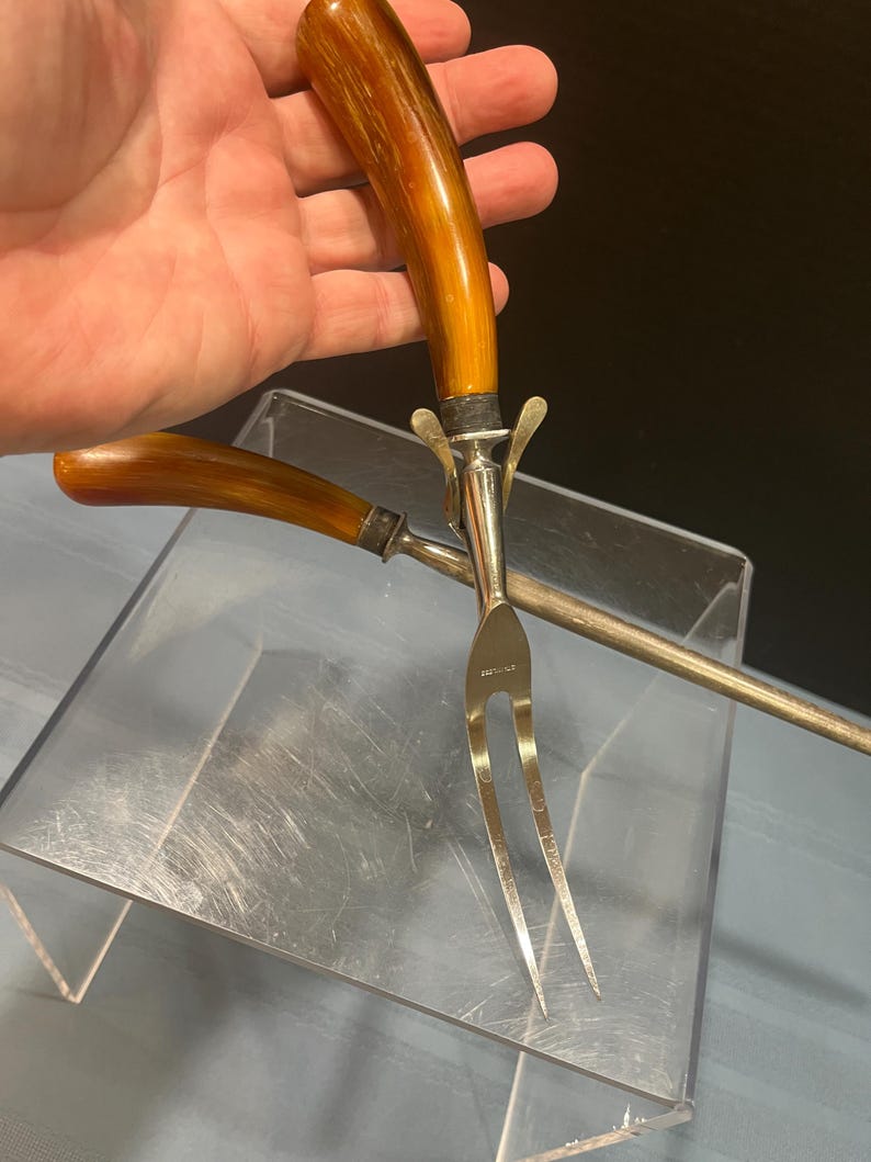 May include: A carving fork and serving fork set with brown handles and silver-colored metal. The carving fork has two tines, and the serving fork has a long, thin handle. The set is displayed on a clear acrylic surface.