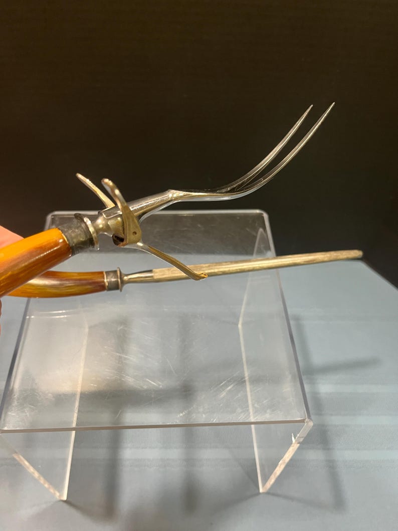 May include: Antique silver-toned carving fork and sharpening steel. The fork has a brown handle and a mechanism to lock the tines. The sharpening steel is long and slender. Both are displayed on a clear acrylic surface.