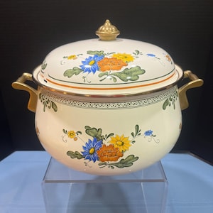 May include: A cream-coloured enamel pot with a matching lid, featuring a floral pattern of blue, orange, and yellow flowers with green leaves. The pot has two gold-toned metal handles on the sides and a gold-toned knob on the centre of the lid.
