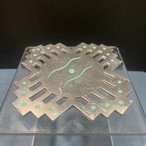 Puede incluir: Un posavasos de metal plateado con un diseño geométrico. El posavasos presenta un diseño circular central con dos líneas verdes curvas y un punto verde. Pequeños puntos verdes se colocan alrededor de los bordes.