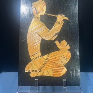 May include: A straw art piece depicting a person playing a flute. The artwork is mounted on a black wooden frame.