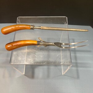 May include: A carving set featuring a sharpening steel and a carving fork. Both tools have wooden handles with a light brown finish and silver-colored metal accents. The fork has two tines.