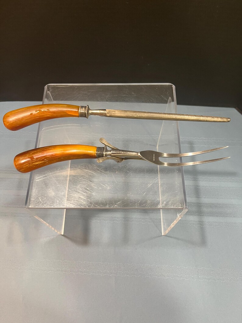 May include: A carving set featuring a sharpening steel and a carving fork. Both tools have wooden handles with a light brown finish and silver-colored metal accents. The fork has two tines.