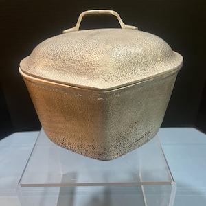 May include: A silver-colored, lidded container with a textured surface. The container has a handle on top and sits on a clear acrylic stand. The container's shape is a rounded square.