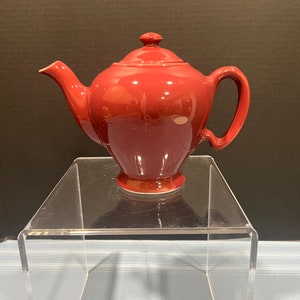 May include: A red ceramic teapot with a lid and a handle. The teapot is sitting on a clear acrylic stand.