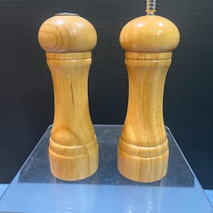 May include: Two wooden salt and pepper grinders. The grinders are made of light-colored wood with a bulbous top and a slender, hourglass-shaped body. The top of one grinder has a silver metal cap. The grinders are displayed on a clear surface.