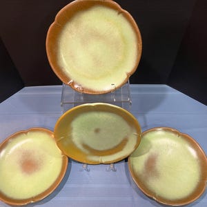 May include: A set of four ceramic plates with a scalloped edge. The plates have a light green center with a brown rim. One plate is oval-shaped, and the other three are round. The plates are displayed on a clear acrylic stand and a blue surface.