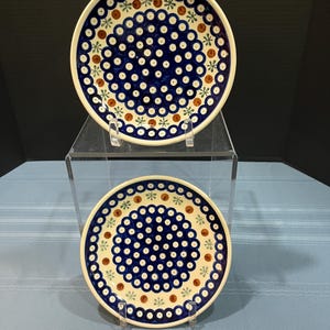 May include: Two ceramic plates with a blue and white floral pattern. The plates have a dark blue center with white dots and are bordered by brown and green accents. The plates are displayed on a clear stand.