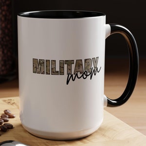 May include: White ceramic mug with a black interior and handle. The mug features the text "MILITARY mom" in a camouflage pattern and black script. The mug is on a wooden surface with a spoon, coffee beans, and a pastry.