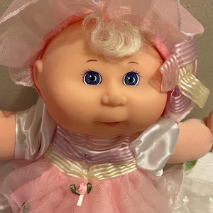 May include: A Cabbage Patch Kids doll with blonde hair, blue eyes, and a soft vinyl face. The doll wears a pink and white dress with a ruffled tulle skirt, a matching bonnet with a striped ribbon bow, and white satin sleeves with pink accents.