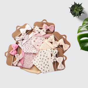 Op de afbeelding: Een verzameling baby slabbetjes en bijpassende haarbanden. De slabbetjes en strikken hebben bloemen- en patroonontwerpen in roze, wit en crème kleuren. De haarbanden hebben zachte, neutraal gekleurde elastische banden. De items worden op bruin karton gepresenteerd.