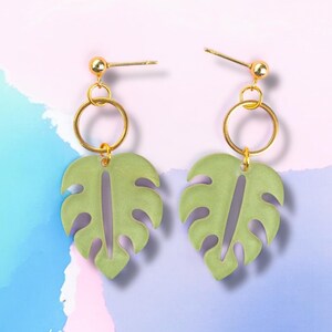 May include: A pair of gold earrings with green monstera leaf charms. The earrings have a simple gold hoop design.