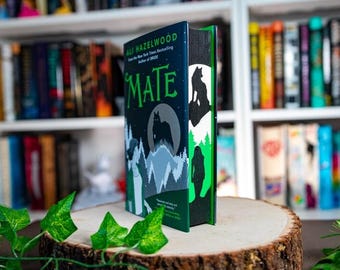 May include: A hardcover book titled "MATE" by Ali Hazelwood, featuring a dark blue cover with a wolf silhouette against a moonlit mountain scene. The book's edges are painted with a green and black forest design. The book is displayed on a wooden slice.