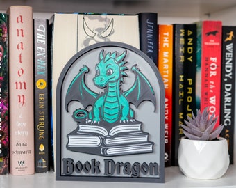 May include: A gray and teal bookend shaped like a dragon holding a stack of books. The bookend says "Book Dragon" on the bottom.