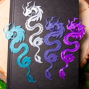 May include: Four dragon-shaped bookmarks in different colors: silver, teal, purple, and dark purple. The bookmarks are on a black surface.