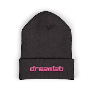 Puede incluir: Un gorro gris oscuro con la palabra "dreamlab" bordada en letras rosas en el borde doblado. El gorro está hecho de un material suave y tejido.