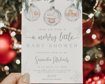 May include: A baby shower invitation card held in front of a Christmas tree. The card features watercolor illustrations of baby items inside ornaments. Text reads "Join us for a merry little baby shower" and event details.