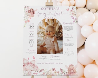 May include: A pink and white first birthday sign with a fairy theme. The sign features a photo of a baby girl, a list of milestones, and the text "Sophia's fairy first year".