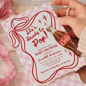 Puede incluir: Una invitaci&oacute;n a un baby shower con una ilustraci&oacute;n de botella de refresco vintage. La invitaci&oacute;n presenta el texto "She's Ready to Pop!" en rojo, junto con los detalles del evento. El fondo tiene un patr&oacute;n de cuadros rosas y blancos.