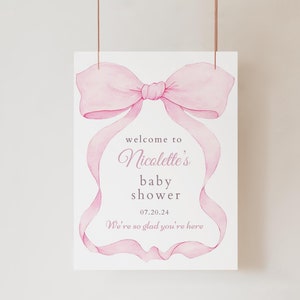 以下が含まれることがあります： 白い背景にピンクの水彩画の弓と「welcome to Nicolette's baby shower 07.20.24 We're so glad you're here」というテキストがあります。