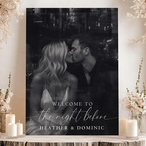 May include: A black and white wedding sign with a couple kissing. The sign reads "WELCOME TO the night Before HEATHER & DOMINIC". The sign is surrounded by white flowers and candles.