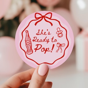 以下が含まれることがあります： 「She's Ready to Pop!」のテキストと赤いアクセントが付いたピンク色の丸いステッカー。ソーダボトル、おしゃぶり、リボンが特徴です。ステッカーはピンク色の風船とギフトボックスの前に保持されています。