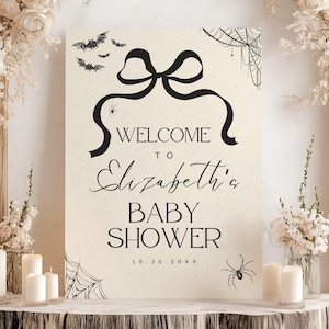 K&ouml;nnte beinhalten: Cremefarbenes Babyparty-Schild mit schwarzen Akzenten. Auf dem Schild steht "Willkommen zu Elizabeths Babyparty" mit dekorativen Flederm&auml;usen, Spinnen und Spinnweben. Das Schild ist mit Blumen und Kerzen dekoriert.