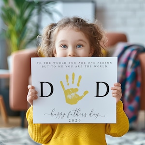 May include: A child holds a white sign with the words "TO THE WORLD YOU ARE ONE PERSON BUT TO ME YOU ARE THE WORLD" and "happy fathers day 2026". The sign has a yellow handprint and the letters "D" on either side.