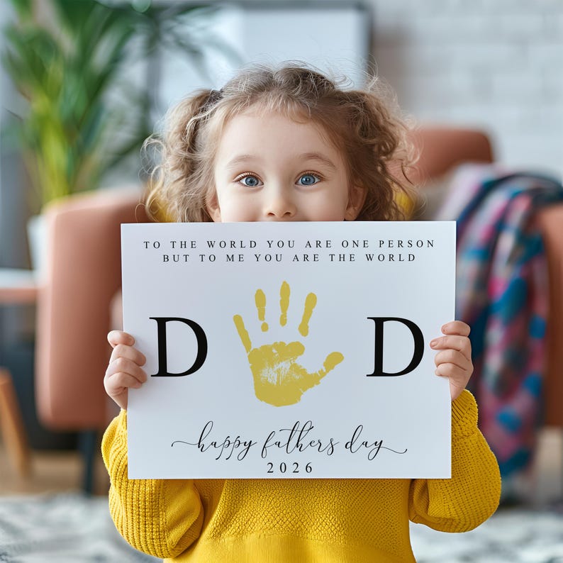 May include: A child holds a white sign with the words "TO THE WORLD YOU ARE ONE PERSON BUT TO ME YOU ARE THE WORLD" and "happy fathers day 2026". The sign has a yellow handprint and the letters "D" on either side.