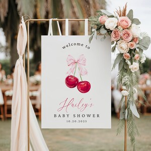 May include: Welcome to Hailey's Baby Shower sign with a pink bow and two red cherries. The sign is on a gold stand with white ribbons.