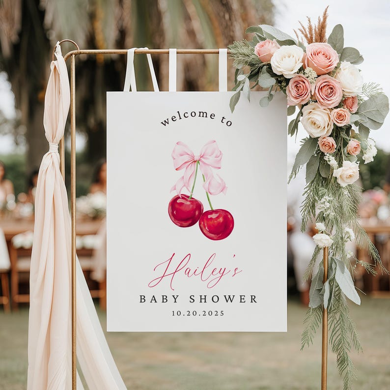 May include: Welcome to Hailey's Baby Shower sign with a pink bow and two red cherries. The sign is on a gold stand with white ribbons.