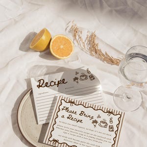 Peut inclure: Nature morte avec des cartes de recettes, une assiette, un verre d'eau et des citrons coup&eacute;s en deux sur un tissu blanc. Les cartes de recettes portent les mentions "Recipe" et "Please Bring a Recipe".