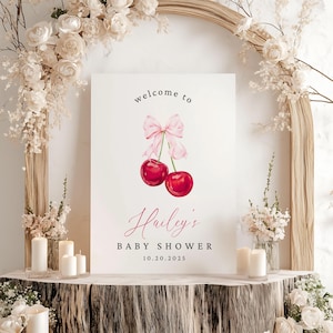 May include: A white welcome sign with a pink bow and two red cherries. The sign reads "Welcome to Hailey's Baby Shower 10.20.2025".