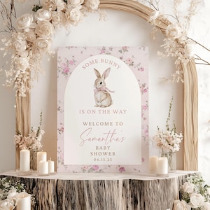 Op de afbeelding: Een roze baby shower-bord met bloemen en een witte boog met een aquarel-illustratie van een bruin konijn met een roze strik. De tekst luidt "Some bunny is on the way. Welcome to Samantha's Baby Shower 04.15.25"