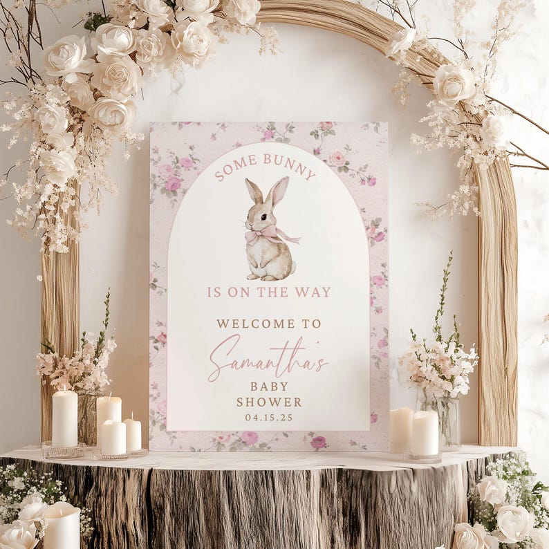 Op de afbeelding: Een roze baby shower-bord met bloemen en een witte boog met een aquarel-illustratie van een bruin konijn met een roze strik. De tekst luidt "Some bunny is on the way. Welcome to Samantha's Baby Shower 04.15.25"