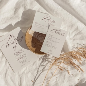 May include: Wedding invitation suite with three cards and a small plate on a white linen surface. The cards include "The Wedding of" and "RSVP" text in elegant script. Dried wheat stalks are in the bottom right corner.