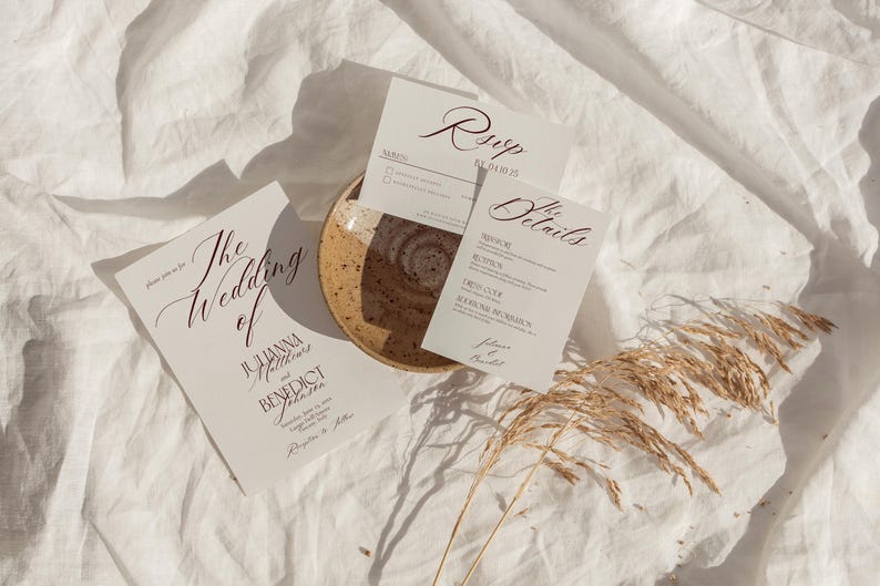 May include: Wedding invitation suite with three cards and a small plate on a white linen surface. The cards include "The Wedding of" and "RSVP" text in elegant script. Dried wheat stalks are in the bottom right corner.