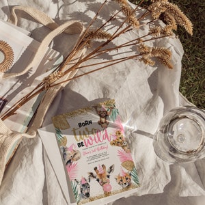 Op de afbeelding: Een verjaardagsuitnodiging met de tekst "Born Two Be Wild" met illustraties van safaridieren. De uitnodiging staat op een witte envelop, met een beige draagtas en gedroogde bloemen op de achtergrond.