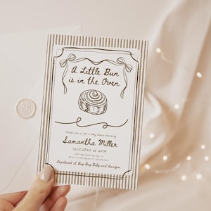 May include: A baby shower invitation with the text "A Little Bun is in the Oven" and a drawing of a cinnamon roll. The invitation is in a decorative frame with a striped background. An envelope with a wax seal is next to the invitation.