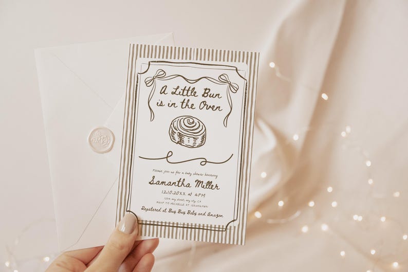 May include: A baby shower invitation with the text "A Little Bun is in the Oven" and a drawing of a cinnamon roll. The invitation is in a decorative frame with a striped background. An envelope with a wax seal is next to the invitation.