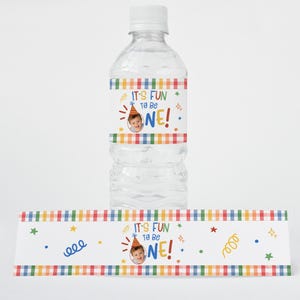 Puede incluir: Una botella de agua de pl&aacute;stico transparente con tap&oacute;n blanco y una etiqueta personalizada con un borde de cuadros de colores, gr&aacute;ficos de confeti y el texto "IT'S FUN TO BE ONE!" junto a una foto circular de un ni&ntilde;o con gorro.