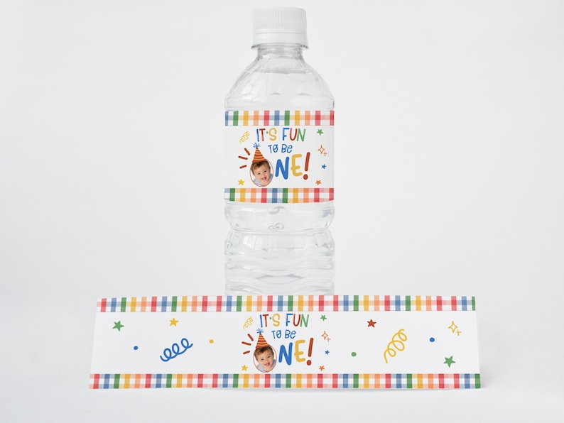 Puede incluir: Una botella de agua de pl&aacute;stico transparente con tap&oacute;n blanco y una etiqueta personalizada con un borde de cuadros de colores, gr&aacute;ficos de confeti y el texto "IT'S FUN TO BE ONE!" junto a una foto circular de un ni&ntilde;o con gorro.