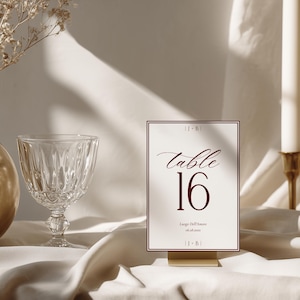 May include: A rectangular table number card featuring the text table 16 in a dark red serif font, with the initials J+B at the top and bottom, and the text Luogo Dell'Amore 06.08.2024 below the number, all framed by a thin dark red border.