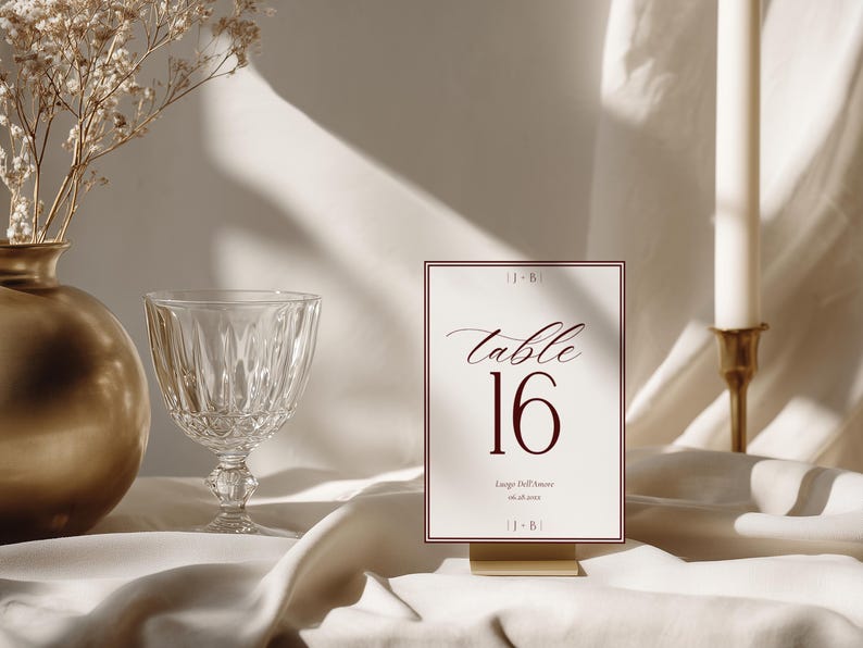 May include: A rectangular table number card featuring the text table 16 in a dark red serif font, with the initials J+B at the top and bottom, and the text Luogo Dell'Amore 06.08.2024 below the number, all framed by a thin dark red border.
