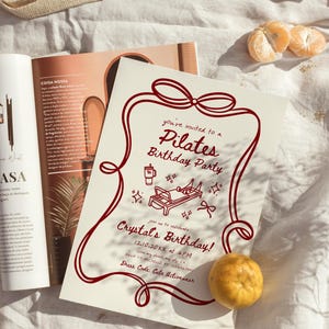 May include: A Pilates birthday party invitation with a burgundy border and text. The invitation includes illustrations of Pilates equipment and a coffee cup. A mandarin orange and a magazine are also visible.