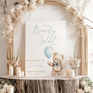 May include: A white sign with the text "We can Bearly Wait" in blue script. Below the text is a cartoon illustration of a brown teddy bear holding a blue balloon. The text "Welcome to Samantha's Baby Shower 12.10.2025" is below the illustration.