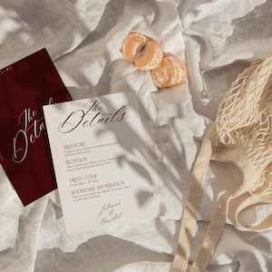 May include: A flat lay featuring wedding details. Two cards with the text "The Details" in elegant script are displayed. One card is burgundy, the other is white. A beige string bag and two tangerine segments are also present.