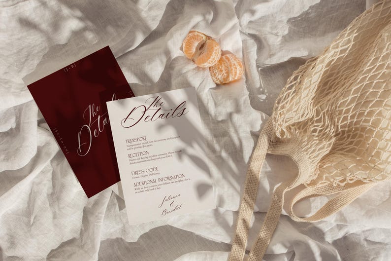 May include: A flat lay featuring wedding details. Two cards with the text "The Details" in elegant script are displayed. One card is burgundy, the other is white. A beige string bag and two tangerine segments are also present.