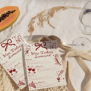 Pu&ograve; includere: Due schede di degustazione di vini con illustrazioni e testo rossi, tra cui un fiocco, calici e bottiglie. Sono visibili anche mezza papaya e un bicchiere d'acqua. Le schede sono in una borsa tote beige.