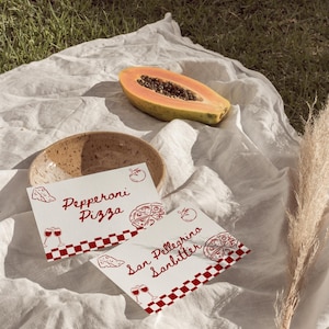 K&ouml;nnte beinhalten: Eine Picknick-Szene mit einem wei&szlig;en Tuch auf Gras. Eine halbierte Papaya, eine Keramikschale und zwei Karten mit rotem Text und Illustrationen sind zu sehen. Auf den Karten steht "Pepperoni Pizza" und "San Pellegrino Sanbitter".